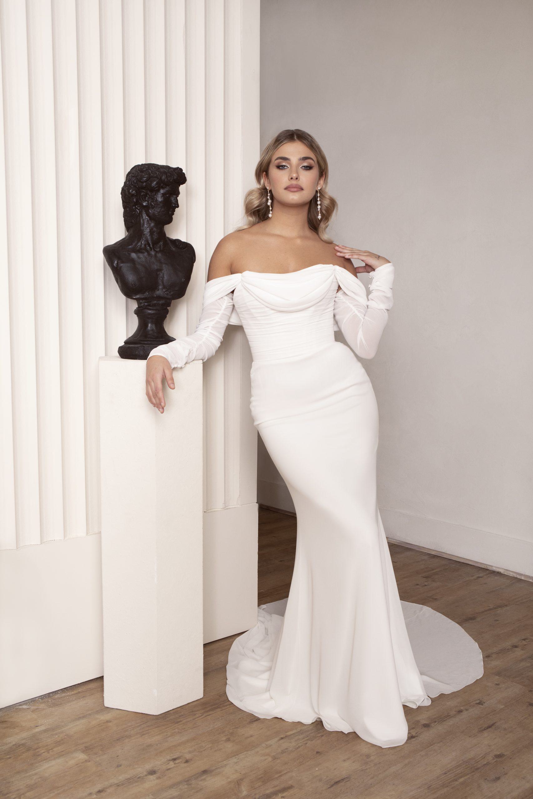 A bride stands confidently beside a pedestal holding a classical bust statue. She is dressed in a sleek, off-the-shoulder wedding gown that hugs her curves, with long fitted sleeves that add an element of classic elegance. The contemporary dress flows smoothly down into a graceful train. Her blonde hair is styled in soft waves, and she holds one hand delicately at her collarbone, creating a poised and refined look. The minimalist setting, with its clean lines and soft lighting, highlights the sophisticated and timeless beauty of the scene.
