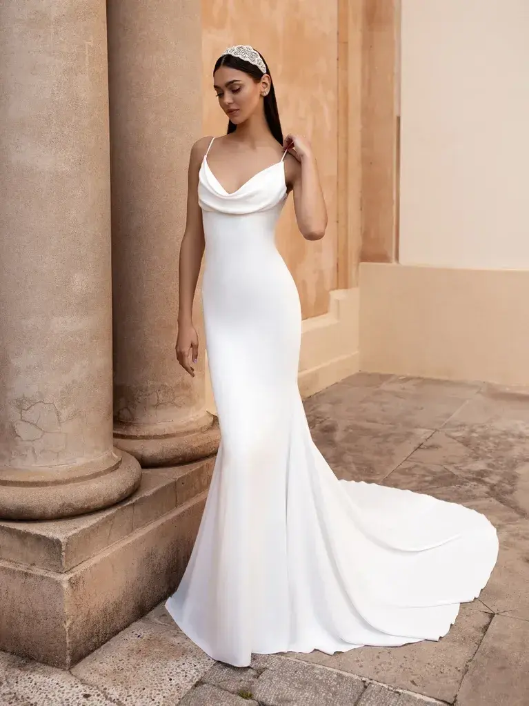 A woman is standing in an elegant architectural setting, wearing a sleek and sophisticated white wedding gown. The gown features thin straps, a cowl neckline, and a form-fitting silhouette that flares out slightly into a train at the bottom. The fabric is smooth and drapes beautifully, emphasizing the simple yet elegant design of the dress. She is positioned near large, classical columns made of stone, with a paved walkway and a warm-toned wall in the background. She is also wearing a headpiece that adds a touch of classic charm to her overall look. The scene conveys a sense of timeless beauty and grace.