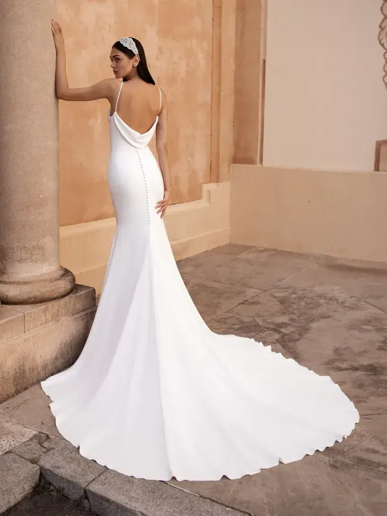 A woman is standing in an elegant architectural setting, wearing a sleek and sophisticated white wedding gown showing the full view of the back and the train. The gown features thin straps, a cowl neckline, and a form-fitting silhouette that flares out slightly into a train at the bottom. The fabric is smooth and drapes beautifully, emphasizing the simple yet elegant design of the dress. She is positioned near large, classical columns made of stone, with a paved walkway and a warm-toned wall in the background. She is also wearing a headpiece that adds a touch of classic charm to her overall look. The scene conveys a sense of timeless beauty and grace.