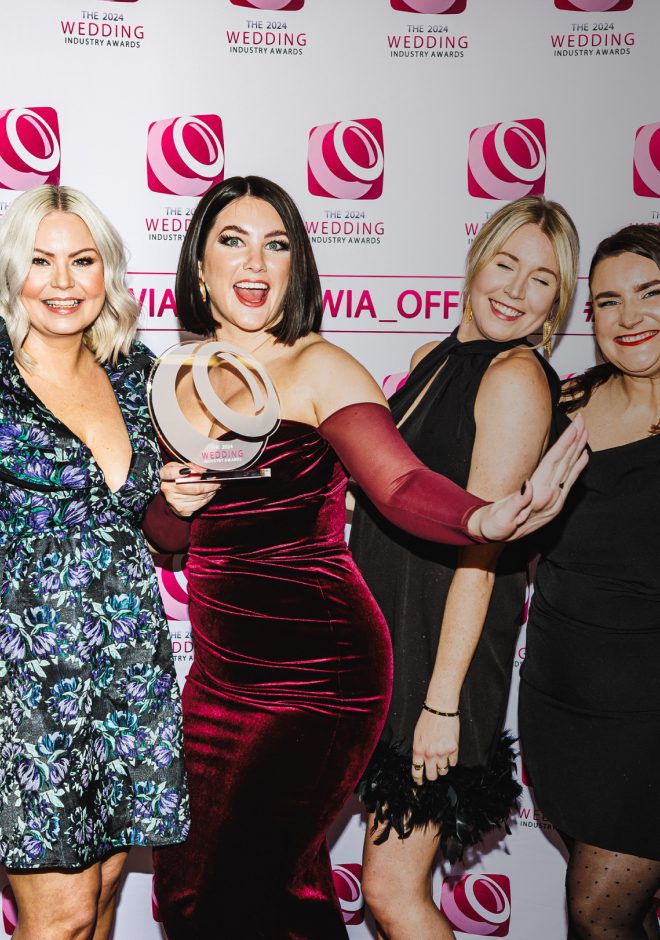 A group of four women stand together, smiling and celebrating. One woman in a maroon velvet dress holds up an award, while the others, dressed in elegant party attire, pose enthusiastically. The background features a pink and white step-and-repeat banner with 'The 2024 Wedding Industry Awards' logo, indicating a celebratory event for wedding industry achievements.