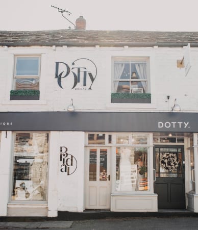 The image shows the exterior of the Dotty bridal boutique. The building is a charming, two-story structure with a white facade. The boutique's name, "Dotty," is prominently displayed in elegant black lettering on the upper portion of the building and above the entrance. There are two large windows on the upper floor, with one of them featuring a small, decorated window box. The ground floor has large display windows on either side of a central entrance door, showcasing bridal gowns and accessories. The boutique's door on the right side has a decorative wreath hanging on it, adding a welcoming touch. The overall appearance of the boutique is stylish and inviting, with a modern yet classic design.