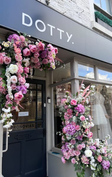 The image shows the entrance of the Dotty bridal boutique, beautifully adorned with a lush and vibrant floral display. The sign above the entrance reads "DOTTY." in sleek white lettering on a dark background. The doorway and window are framed with an abundance of pink, purple, and white flowers, creating a stunning and inviting entrance. The large display window next to the door showcases elegant bridal gowns. The floral arrangement adds a touch of romance and charm to the boutique's exterior, making it an eye-catching and welcoming sight for visitors.