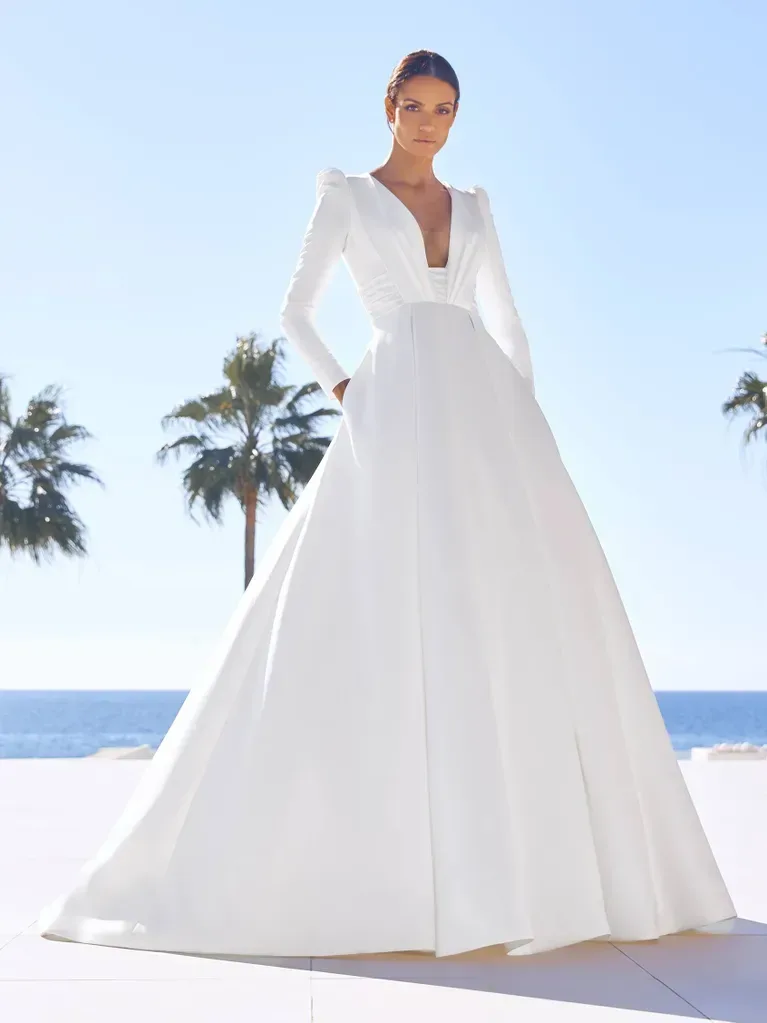 A woman is standing outdoors in a bright, sunny setting, wearing an elegant long-sleeved white ball gown. The gown features a deep V-neckline and a fitted bodice that extends into a full, voluminous skirt. The fabric is smooth and structured, creating a classic and regal silhouette. The background shows a clear blue sky with palm trees and a view of the ocean, suggesting a tropical or coastal location. The bright sunlight enhances the pristine whiteness of the gown and adds a sense of freshness and luxury to the scene.
