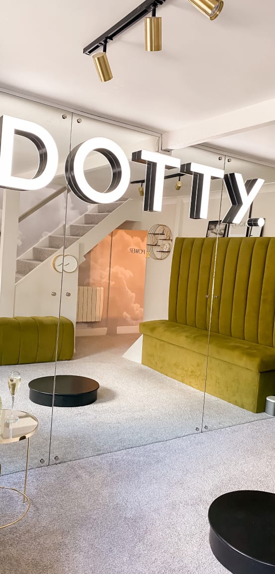 The image shows the interior of the Dotty bridal boutique, focusing on a stylish seating area. The space features a large green velvet bench with high backs, providing a luxurious and comfortable place for clients to sit. In front of the bench, there are two small round black tables. The floor is covered with a plush, light coloured carpet. Above the seating area, the word "DOTTY" is prominently displayed in large white letters suspended from the ceiling. In the background, there is a staircase and some decorative elements on the walls, adding to the boutique's chic and modern ambiance. The overall setting is designed to be elegant and inviting, perfect for bridal fittings and consultations.