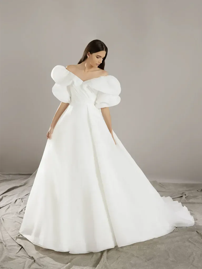 A woman is standing in a minimalist studio setting, wearing a dramatic white ball gown. The gown features an off-the-shoulder design with large, voluminous ruffled sleeves and a fitted bodice that flares out into a full, floor-length skirt. The fabric is smooth and structured, giving the dress a bold and elegant silhouette. The background is plain, with draped cloth on the floor, creating a neutral and understated environment that highlights the striking design and grandeur of the gown.