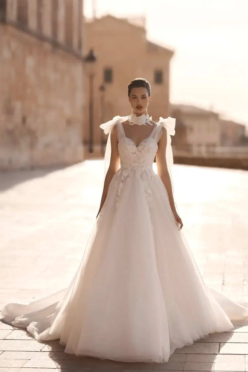The image shows a bride wearing a stunning white wedding dress with a high neckline and intricate lace detailing on the bodice. The dress features a fitted waist and flows into a voluminous, full-length skirt with a long, elegant train that trails behind her. The bodice includes delicate, sheer lace panels, adding a romantic and sophisticated touch to the gown. The bride's hair is styled in a sleek updo, and she is adorned with a sheer veil that cascades from her shoulders, further enhancing the ethereal and graceful appearance. Her pose is poised and elegant, capturing the timeless beauty of the gown.