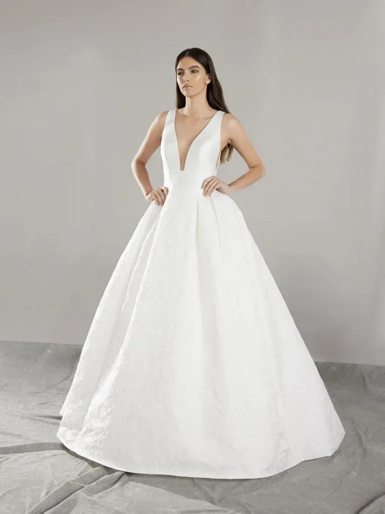 A woman is standing in a minimalist studio setting, wearing a voluminous white ball gown. The gown features a deep V-neckline and sleeveless design, with a fitted bodice that flows into a full, floor-length skirt. The fabric of the dress is smooth and structured, creating a classic and elegant silhouette. The backdrop is plain, with draped cloth on the floor, providing a neutral and understated environment that emphasizes the grandeur of the dress.