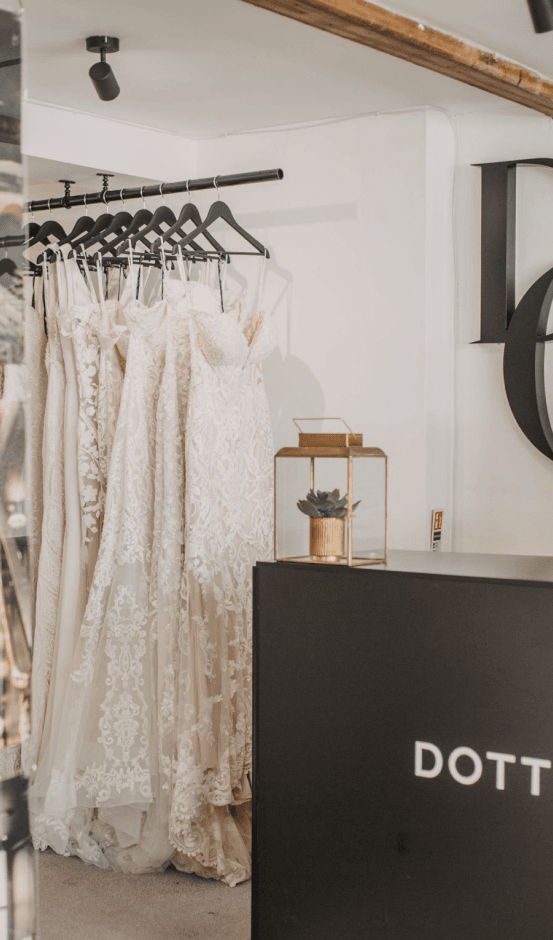 The image shows the interior of the Dotty bridal boutique. In the foreground, there is a sleek, black reception desk with the word "DOTTY." written on it in white letters. Behind the desk, there is a small glass container with a succulent plant inside, adding a touch of greenery to the decor. To the left of the desk, several wedding dresses are hanging on a rack, showcasing various designs and fabrics. The background features a modern and minimalist aesthetic with a white wall and a large, decorative "Dotty" logo partially visible. The overall atmosphere of the boutique is elegant and inviting, highlighting the beautiful bridal gowns on display.