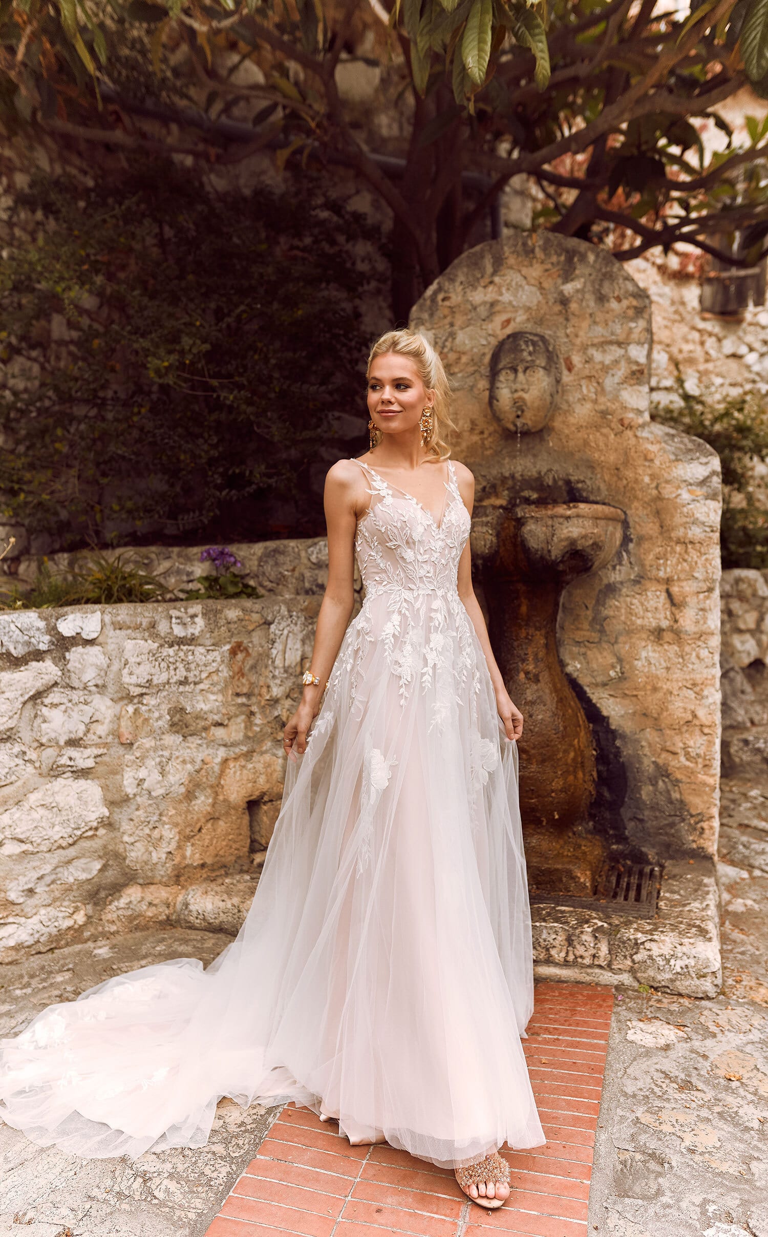 A woman is wearing a beautiful wedding dress with a fitted bodice adorned with intricate lace detailing. The dress features thin, delicate straps and a flowing, ethereal skirt that extends into a long, elegant train. The overall design is romantic and graceful, emphasizing the woman's slender figure and adding a touch of fairy-tale elegance to her look. Her blonde hair is styled in soft waves, complementing the delicate and refined aesthetic of the dress.