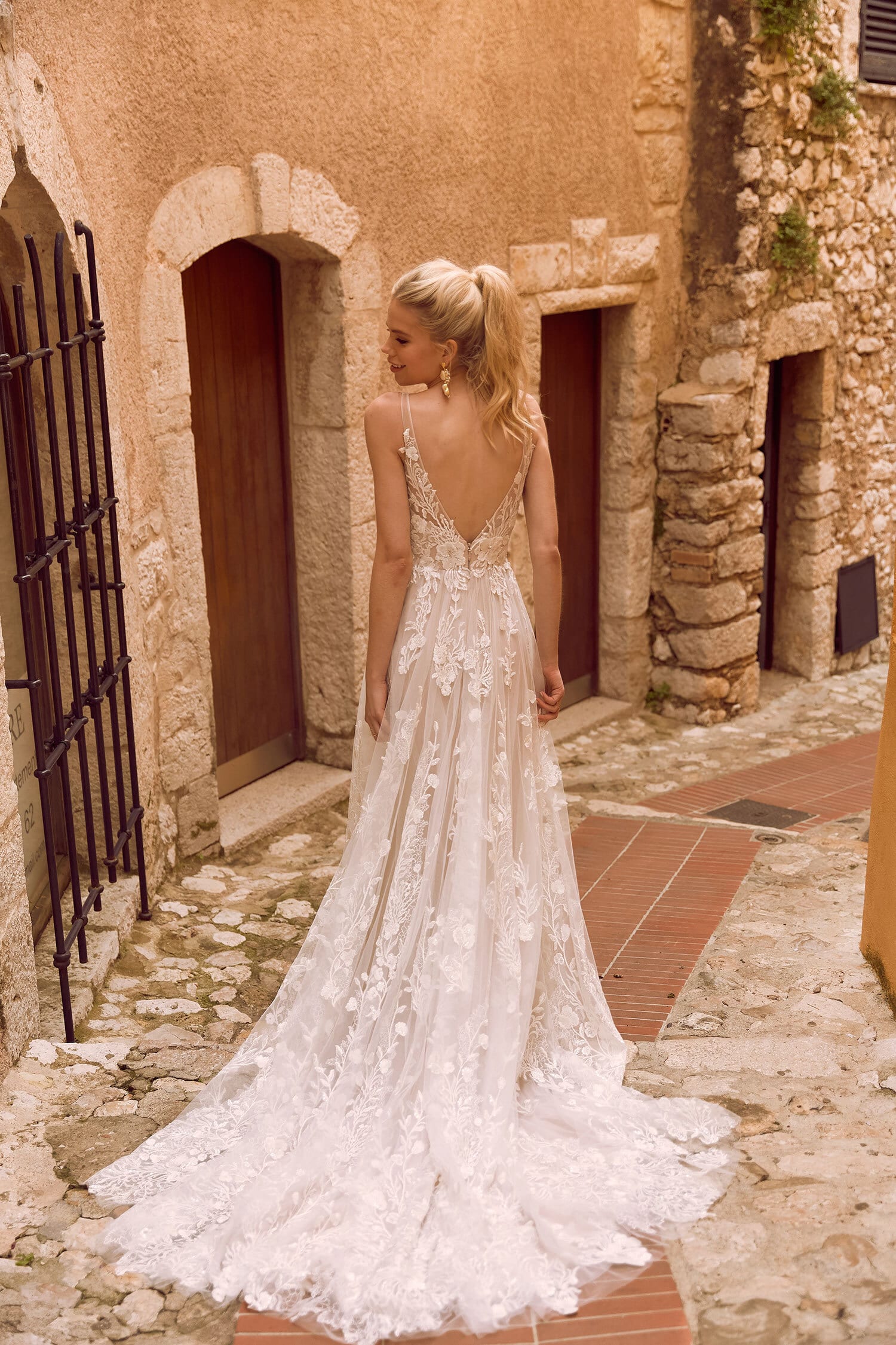 The image shows a woman wearing an elegant and romantic white wedding gown viewed from the back. The dress features a deep V-back neckline with delicate lace detailing that extends to the bodice and straps, creating a refined and feminine look. The bodice is fitted, accentuating the woman's figure, and transitions into a flowing, A-line skirt. The skirt is made of light, airy fabric adorned with intricate lace patterns, adding texture and visual interest to the gown. The woman's hair is styled in soft waves, cascading down her back, complementing the graceful and ethereal design of the dress. The overall effect is one of timeless beauty and romantic elegance, perfect for a fairy-tale wedding.