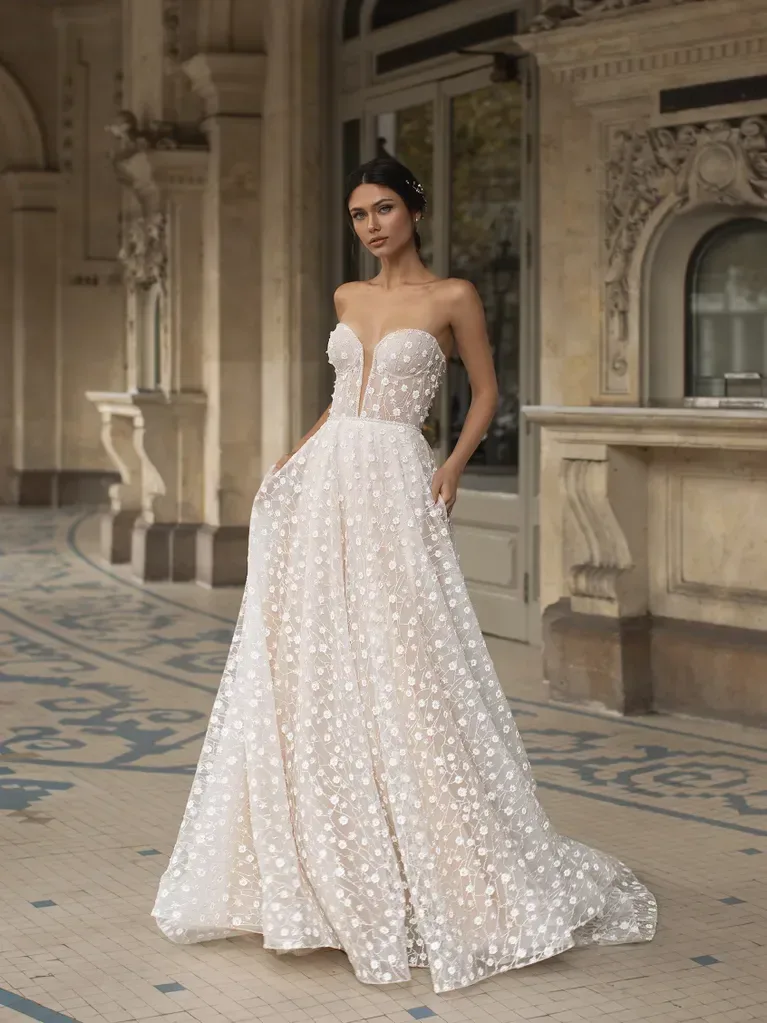 A woman is standing in an ornate, historical architectural setting, wearing a strapless white ball gown. The gown features a deep sweetheart neckline and is adorned with an intricate pattern of small, delicate floral appliqués. The fabric is textured and creates a voluminous, floor-length skirt that adds to the elegance of the dress. The background includes detailed stone carvings, arches, and a tiled floor, enhancing the luxurious and regal atmosphere of the scene. The overall ambiance is one of timeless sophistication and elegance.