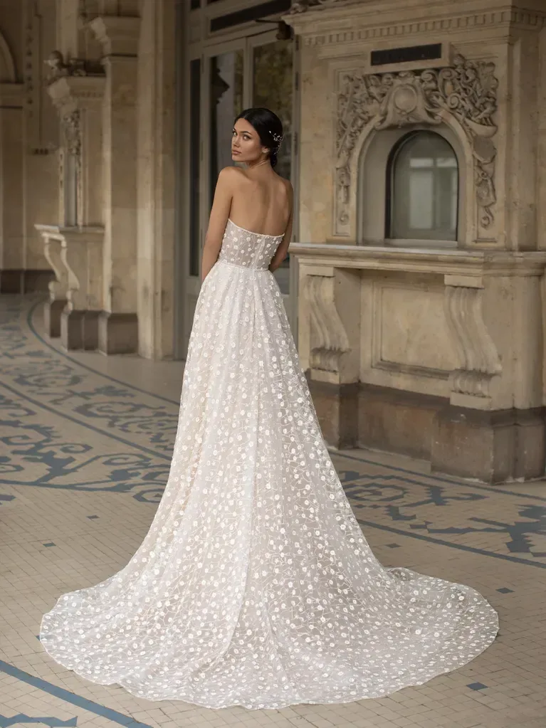 A woman is standing in an ornate, historical architectural setting, wearing a strapless white ball gown shown here the full back and train. The gown features a deep sweetheart neckline and is adorned with an intricate pattern of small, delicate floral appliqués. The fabric is textured and creates a voluminous, floor-length skirt that adds to the elegance of the dress. The background includes detailed stone carvings, arches, and a tiled floor, enhancing the luxurious and regal atmosphere of the scene. The overall ambiance is one of timeless sophistication and elegance.