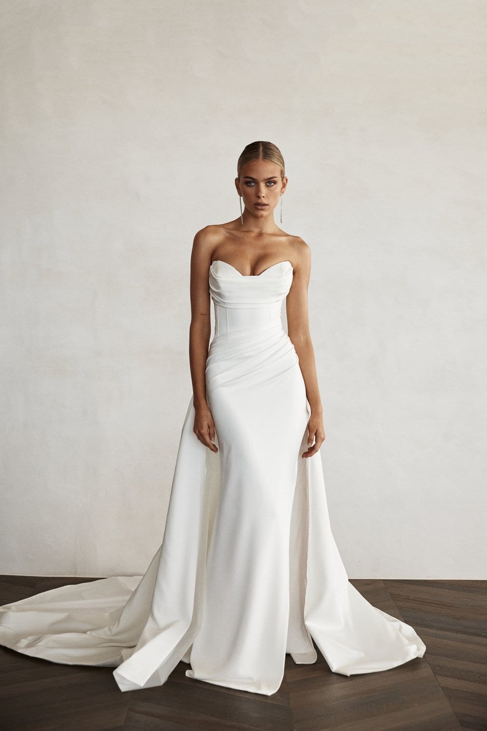 The image shows a woman wearing a white, strapless wedding dress with a sweetheart neckline. The dress is fitted through the bodice and hips, flaring out into a flowing train that spreads elegantly on the floor. The woman stands with a neutral expression, her arms relaxed at her sides. The background is simple and plain, with a light-colored wall and a dark floor, drawing all attention to the dress and the wearer.