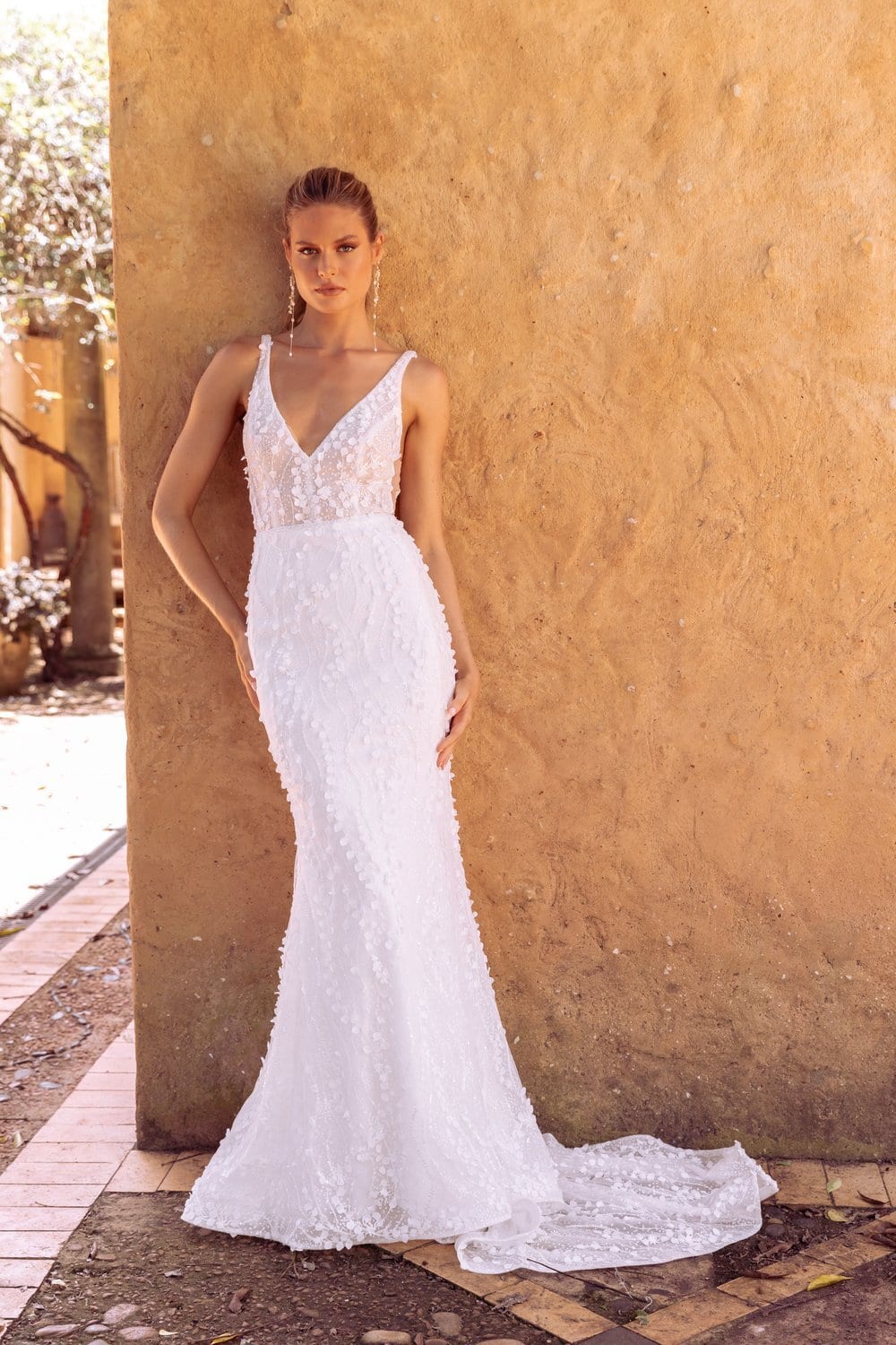 The image shows a woman wearing a white, sleeveless wedding dress with a deep V-neckline and lace detailing on the bodice. The dress is fitted through the torso and hips, flaring out slightly into a small train that drapes elegantly on the tiled floor. The woman is standing outdoors, leaning against a textured, tan-colored wall with one hand resting on it and the other by her side. The background includes a paved area with a glimpse of greenery and another structure in the distance, suggesting a rustic or Mediterranean setting.