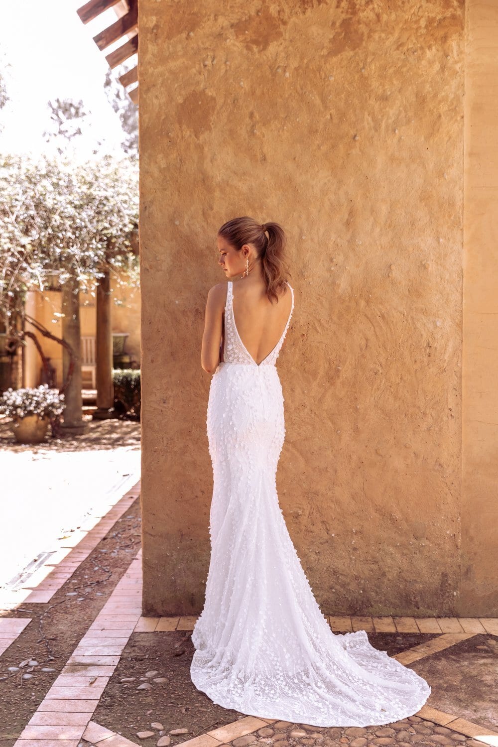 The image shows a woman wearing a white, sleeveless wedding dress with a deep V-neckline and lace detailing on the bodice. Showing the back view with deep square backline. The dress is fitted through the torso and hips, flaring out slightly into a small train that drapes elegantly on the tiled floor. The woman is standing outdoors, leaning against a textured, tan-colored wall with one hand resting on it and the other by her side. The background includes a paved area with a glimpse of greenery and another structure in the distance, suggesting a rustic or Mediterranean setting.