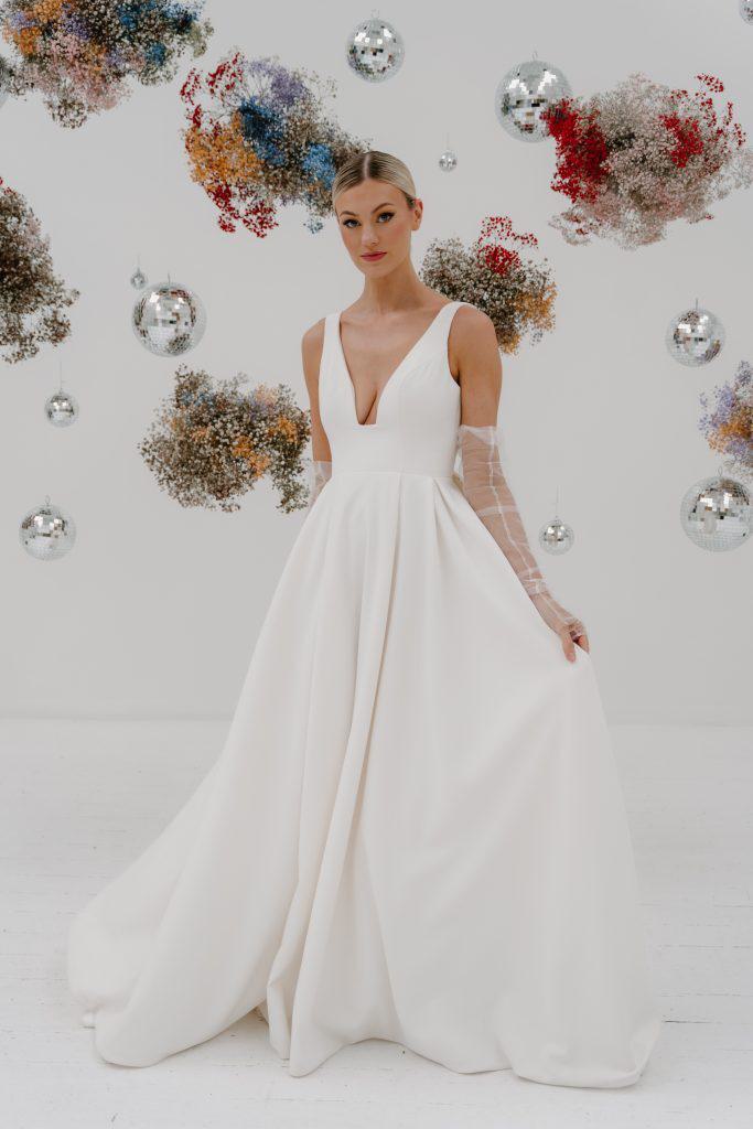 A classic A-line, contemporary wedding dress featuring a deep V-neckline and a fitted bodice. The dress flows into a voluminous, full skirt with a smooth, elegant finish, complemented by sheer, long sleeves adorned with delicate appliqués.