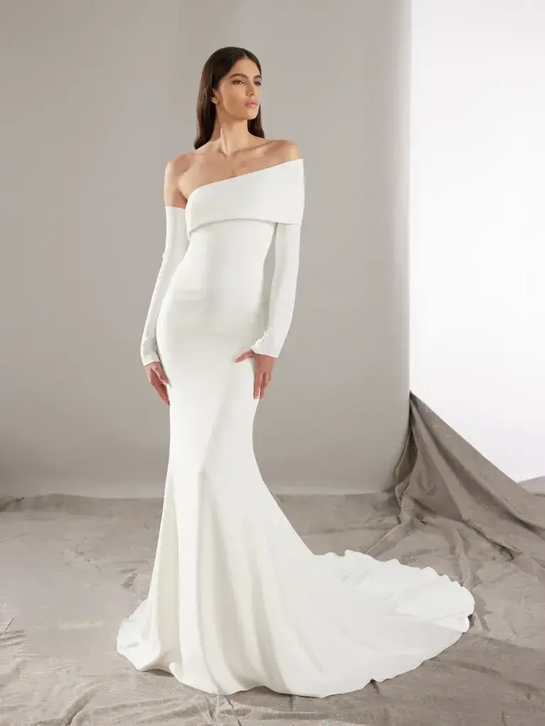 A woman is standing in a minimalist studio setting, wearing a sophisticated off-the-shoulder white gown. The gown is form-fitting with long sleeves and a sleek silhouette that flares out into a small train at the bottom. The fabric is smooth and elegant, emphasizing the clean lines and simplicity of the design. The backdrop is plain, with draped cloth on the floor, creating a neutral and understated environment that highlights the elegance of the dress.