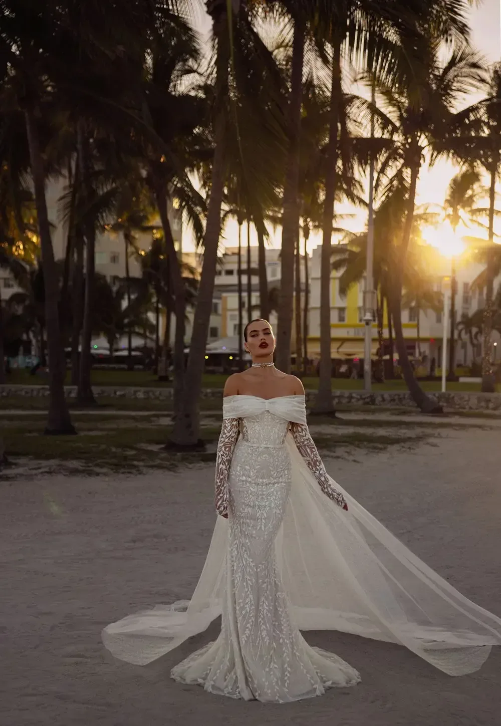 The image shows a person wearing an elegant, off-the-shoulder white wedding gown. The gown features long, fitted sleeves that extend to the wrists and a form-fitting bodice that accentuates the figure. The dress flares out at the bottom into a mermaid-style skirt with a long, flowing train that adds a dramatic effect. The person also wears a headpiece that complements the sophisticated and glamorous look of the gown.