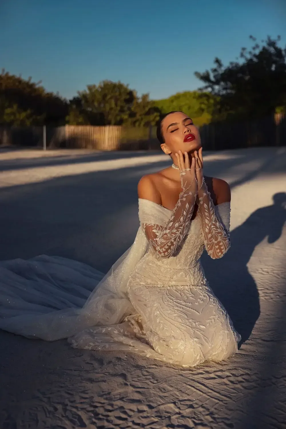 The image shows a woman wearing an elegant off-the-shoulder wedding dress, kneeling in a graceful pose. The gown features long, fitted lace sleeves that extend to the wrists and a form-fitting bodice adorned with intricate lace details. The dress flows into a skirt that drapes softly around her. The woman's pose is expressive, with her hands gently touching her face, and she has an elegant, contemplative expression. The overall look is sophisticated and romantic, highlighting the intricate details and the elegance of the gown.