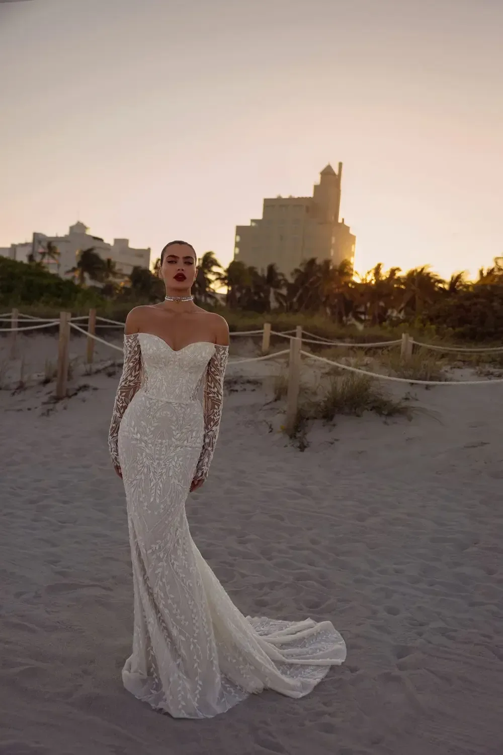 The image shows a bride wearing an off-the-shoulder, white wedding dress. The gown features long, fitted sleeves made of intricate lace that reach to the wrists. The bodice is form-fitting and leads into a mermaid-style skirt that flares out at the bottom. The dress includes a short, elegant train. The overall look is sophisticated and glamorous, highlighting the bride's figure beautifully.