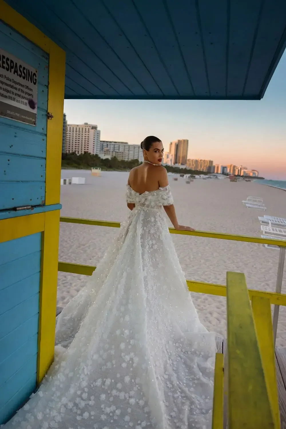The Wona Miami Bliss Couture Maryland 3 gown is an exquisite off-the-shoulder dress that exudes elegance and sophistication. The bodice features delicate floral appliqués, flowing seamlessly into a voluminous skirt adorned with subtle sequins and intricate detailing. The gown's train cascades beautifully, creating a breathtaking silhouette. The bride, with her hair styled in a sleek updo, gazes confidently towards the horizon, framed by the vibrant blue and yellow lifeguard station. The serene beach backdrop at sunset, with its soft sand and distant cityscape, enhances the gown's timeless beauty, making this a truly stunning bridal portrait.