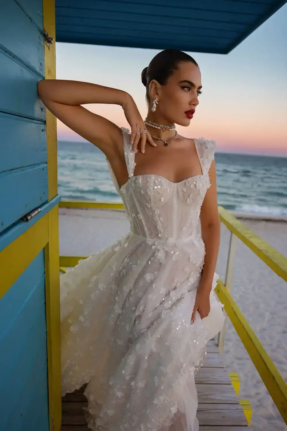 The Wona Miami Bliss Couture Maryland 4 gown is a breathtaking masterpiece, featuring a structured corset bodice adorned with delicate floral appliqués and subtle sequins that catch the light beautifully. The gown's intricate design is accentuated by its sweetheart neckline and elegant straps, offering a romantic yet modern silhouette. The bride's sleek, pulled-back hairstyle and bold red lipstick enhance the gown's sophisticated allure. Set against the backdrop of a serene beach at sunset, the vibrant blue and yellow lifeguard station adds a touch of playful contrast to the timeless elegance of the dress.