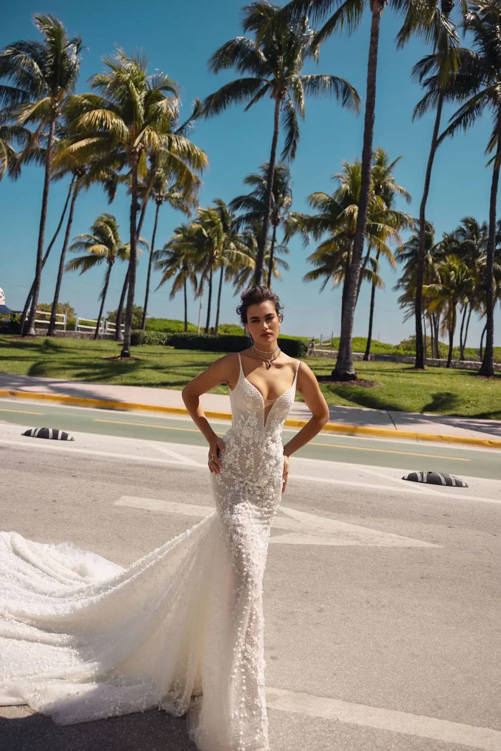 A bride poses confidently on a sunny Miami street, wearing the Wona Miami Bliss Couture Tennessee 1 gown. The dress features a deep plunging neckline and delicate spaghetti straps, emphasizing her figure. The intricate lace detailing covers the dress, showcasing a floral pattern that flows seamlessly into a dramatic, elegant train. Her hair is styled in a chic updo, and she wears a statement necklace that complements the gown’s sophisticated design. The background of palm trees and clear blue sky adds a vibrant and tropical touch, highlighting the bride’s modern elegance and the dress’s exquisite craftsmanship.