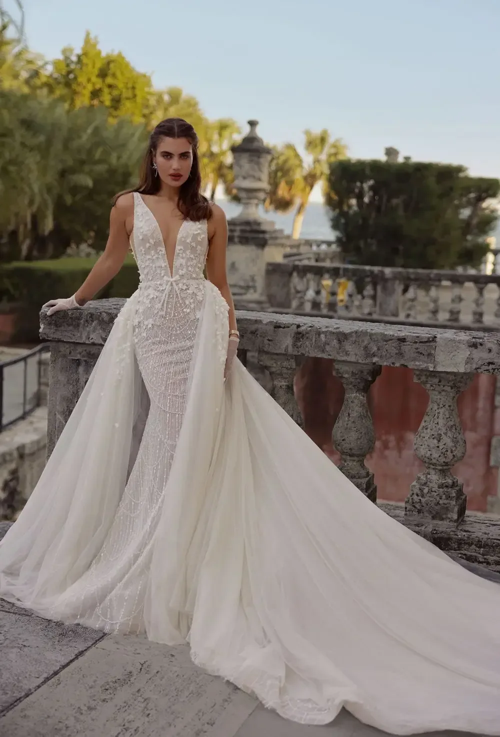 A model poses in the Wona Miami Bliss Couture Wisconsin 1 gown, standing on an elegant stone terrace with lush greenery and classic architectural elements in the background. The gown features a deep V-neckline and intricate beadwork on the bodice, complemented by a flowing overskirt that adds volume and drama to the silhouette. The model's hair is styled in loose waves, and she accessorizes with statement earrings and delicate gloves. The picturesque setting, combined with the gown's stunning details, creates a sophisticated and romantic atmosphere.
