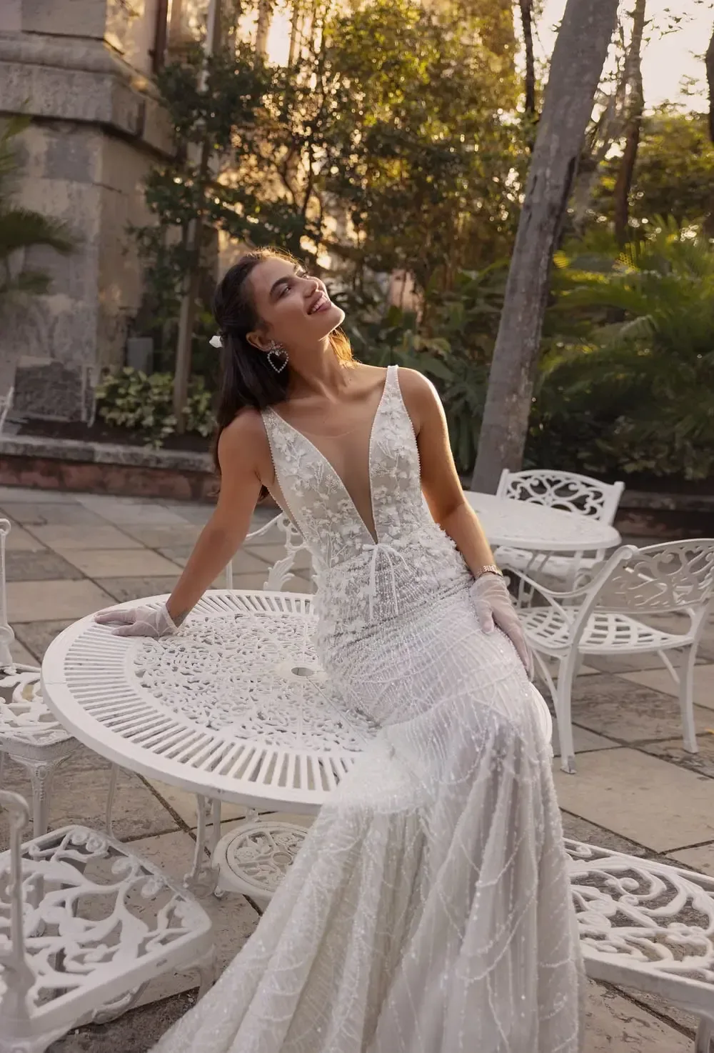 A model wearing the Wona Miami Bliss Couture Wisconsin 2 gown poses in an outdoor setting with white wrought-iron furniture. The gown features a deep V-neckline and is adorned with intricate beadwork and lace details, creating a glamorous and elegant look. The model's hair is styled with soft waves, and she accessorizes with statement earrings and delicate gloves. The evening sunlight enhances the scene, casting a warm glow on the model and highlighting the gown's exquisite details. The lush greenery and vintage furniture add to the romantic and sophisticated ambiance.